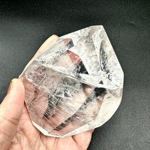 May include: A large, clear quartz crystal with a faceted, geometric shape. The crystal is held in a hand, showcasing its transparency and internal patterns. The crystal's surface reflects light, creating a sparkling effect.