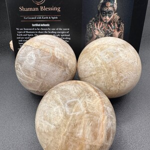 Pode incluir: Tr&ecirc;s esferas de selenita bege com uma textura cristalina branca. As esferas est&atilde;o dispostas em uma forma&ccedil;&atilde;o triangular sobre uma superf&iacute;cie cinza escura. O texto "Shaman Blessing" &eacute; vis&iacute;vel no fundo.