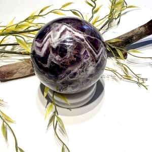 Pu&ograve; includere: Una grande sfera di ametista viola con vortici bianchi e marroni. La sfera &egrave; appoggiata su un piedistallo bianco.