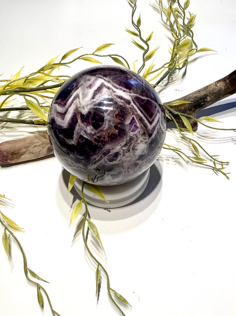 Pu&ograve; includere: Una grande sfera di ametista viola con vortici bianchi e marroni. La sfera &egrave; appoggiata su un piedistallo bianco.