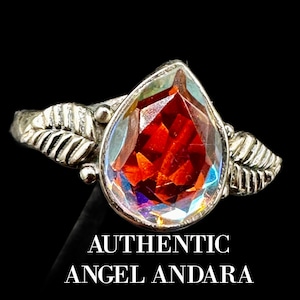 Puede incluir: Un anillo de plata con una piedra preciosa roja e iridiscente en forma de lágrima engastada en el centro. La banda presenta dos diseños en forma de hoja a cada lado de la piedra. El texto "AUTHENTIC ANGEL ANDARA" está impreso debajo del anillo.