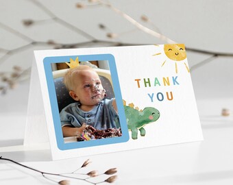 May include: A white thank you card with a blue border and a photo of a child wearing a crown and covered in cake. The card also features a green dinosaur and a yellow sun with a smiling face. The text "THANK YOU" is written in colorful letters.
