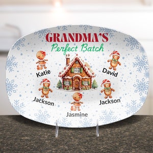 May include: White oval ceramic serving platter with a gingerbread house and gingerbread people. The platter has a blue snowflake pattern and the text "Grandma's Perfect Batch". The gingerbread people are labeled "Katie", "Jackson", "David", "Jackson", and "Jasmine".