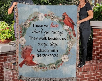Könnte beinhalten: Eine gewebte Wolldecke mit blauem Hintergrund und einem Kranz aus Kiefernästen, Baumwollknäueln und roten Kardinälen. Der Text lautet "Those we love don't go away They walk besides us every day Chad Smith 04/20/2024".