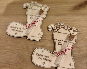 May include: Two wooden Christmas stocking ornaments with a test tube and cork. The ornaments have the text "Kleiner Gruß vom Nikolaus". Each test tube is tied with red and white twine. The stockings are decorated with stars and candy canes.