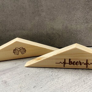 Könnte beinhalten: Zwei Bier-Untersetzer aus Holz. Ein Untersetzer zeigt ein geätztes Design von zwei anstoßenden Bierkrügen. Der andere Untersetzer hat das Wort "beer" mit einer Herzschlaglinie. Beide sind helles Holz.