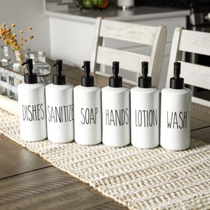 May include: Six white ceramic pump bottles with black pumps, labeled "DISHE", "SANITIZE", "SOAP", "HANDS", "LOTION", and "WASH" in black text. The bottles are arranged on a beige woven table runner, with a wooden table and chairs in the background.