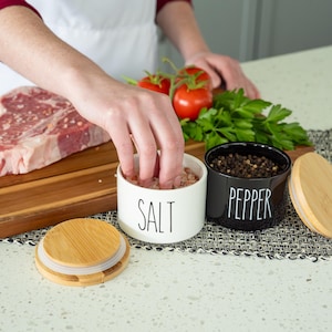 May include: White ceramic salt shaker with a black label that says "SALT" and a black ceramic pepper shaker with a black label that says "PEPPER". Both shakers have bamboo lids.