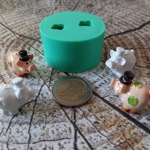 Könnte beinhalten: Vier kleine Sparschweine-Figuren mit schwarzen Zylindern, zwei weiße und zwei rosa, eine mit grünen Kleeblatt-Akzenten, auf einer Holzoberfläche mit einer grünen Silikonform zum Herstellen von Sparschweinen.