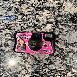 May include: A disposable camera with a pink and white cow print pattern. The front features a photograph of a person, the text "Kay Kay", "Turn on flash", and "Houston 2025". The camera includes a central lens, a viewfinder, and a flash unit.