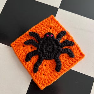 Op de afbeelding: Een vierkant gehaakt granny square in feloranje garen met een zwarte gehaakte spin in het midden. De spin heeft acht getextureerde poten en twee kleine, opvallende roze geborduurde ogen op de ronde zwarte kop van het spinnenfiguur.