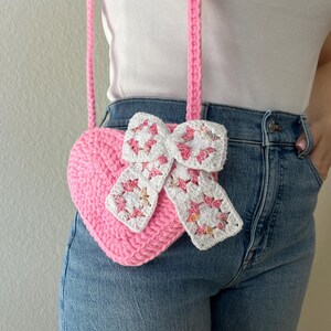 May include: A pink crocheted heart-shaped crossbody bag with a white and pink bow detail. The bag has a long pink strap. The bag is handmade and has a textured appearance. The bag is worn over a white top and blue jeans.
