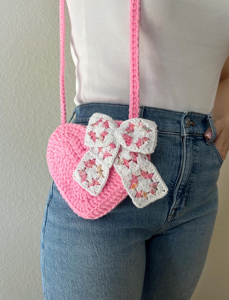 May include: A pink crocheted heart-shaped crossbody bag with a white and pink bow detail. The bag has a long pink strap. The bag is handmade and has a textured appearance. The bag is worn over a white top and blue jeans.