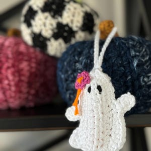 Op de afbeelding: Een wit gehaakt spookornament met zwarte geborduurde ogen en een kleine roze bloem met een oranje steeltje aan de zijkant. Het spookje hangt aan een wit garenlusje voor drie gestructureerde, ronde, gehaakte pompoenen in donkerblauw, roze en zwart-wit.