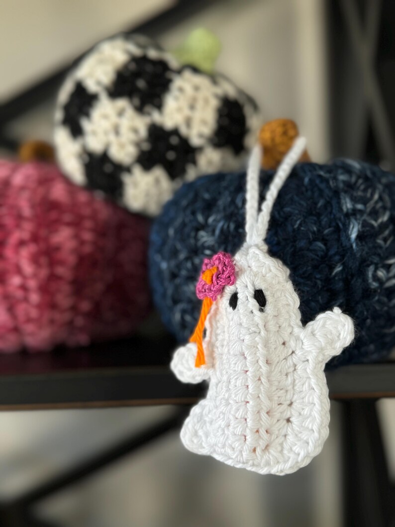Op de afbeelding: Een wit gehaakt spookornament met zwarte geborduurde ogen en een kleine roze bloem met een oranje steeltje aan de zijkant. Het spookje hangt aan een wit garenlusje voor drie gestructureerde, ronde, gehaakte pompoenen in donkerblauw, roze en zwart-wit.