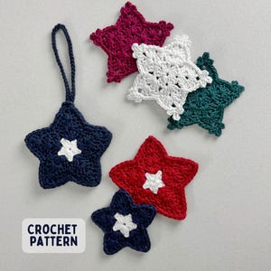 May include: Five star-shaped crochet ornaments in dark blue, red, white, dark green, and magenta, each featuring a small white star center. The dark blue star includes a hanging loop. A white rectangular label displays the text CROCHET PATTERN.
