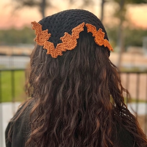 May include: A black crochet bandana with an orange bat design around the edge. The bandana is worn on the head of a person with long, curly brown hair.