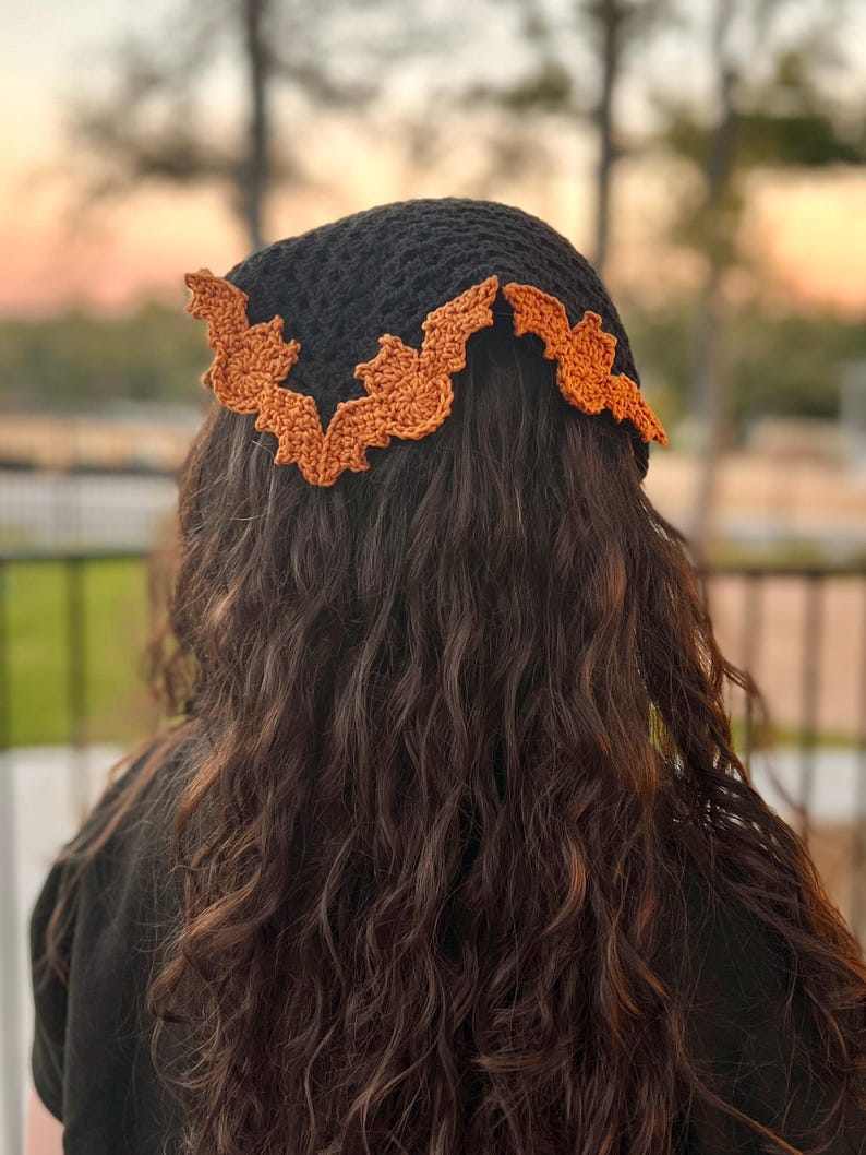 May include: A black crochet bandana with an orange bat design around the edge. The bandana is worn on the head of a person with long, curly brown hair.