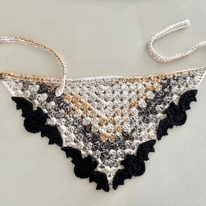 May include: A crocheted triangle scarf with a black and white bat design. The scarf is made with a variety of beige, brown, and black yarns.