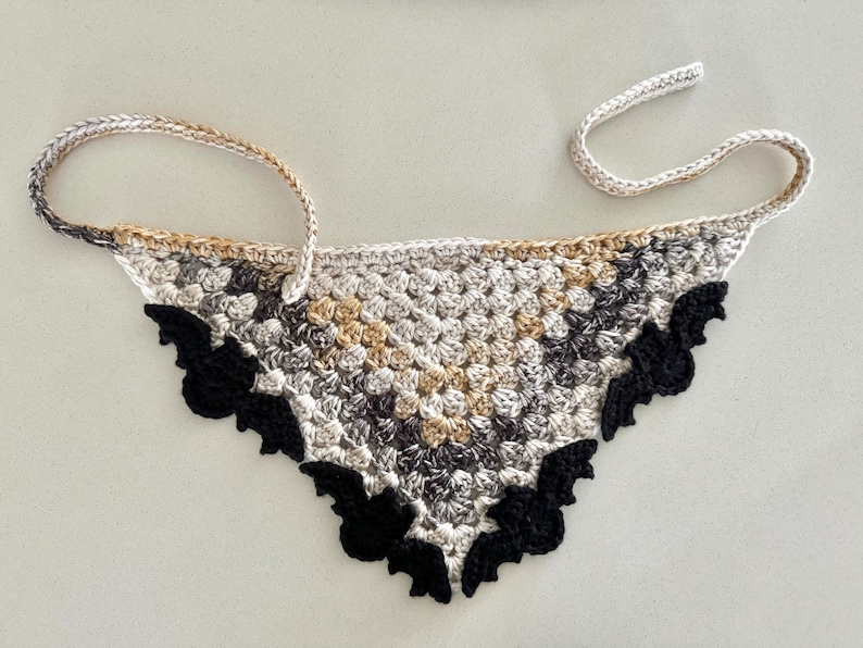 May include: A crocheted triangle scarf with a black and white bat design. The scarf is made with a variety of beige, brown, and black yarns.