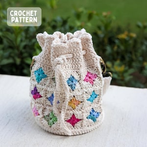 Peut inclure: Sac en crochet crème avec des motifs en forme d'étoiles colorées. Le sac a une fermeture à cordon et une poignée. Le texte "CROCHET PATTERN" est dans un encadré blanc. Le sac est posé sur une surface blanche.