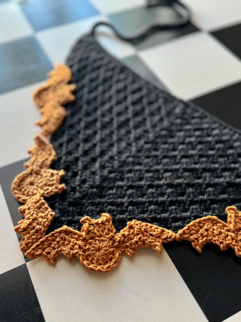 May include: A black crocheted scarf with a brown crocheted bat design around the edge. The scarf is lying on a black and white checkered surface.