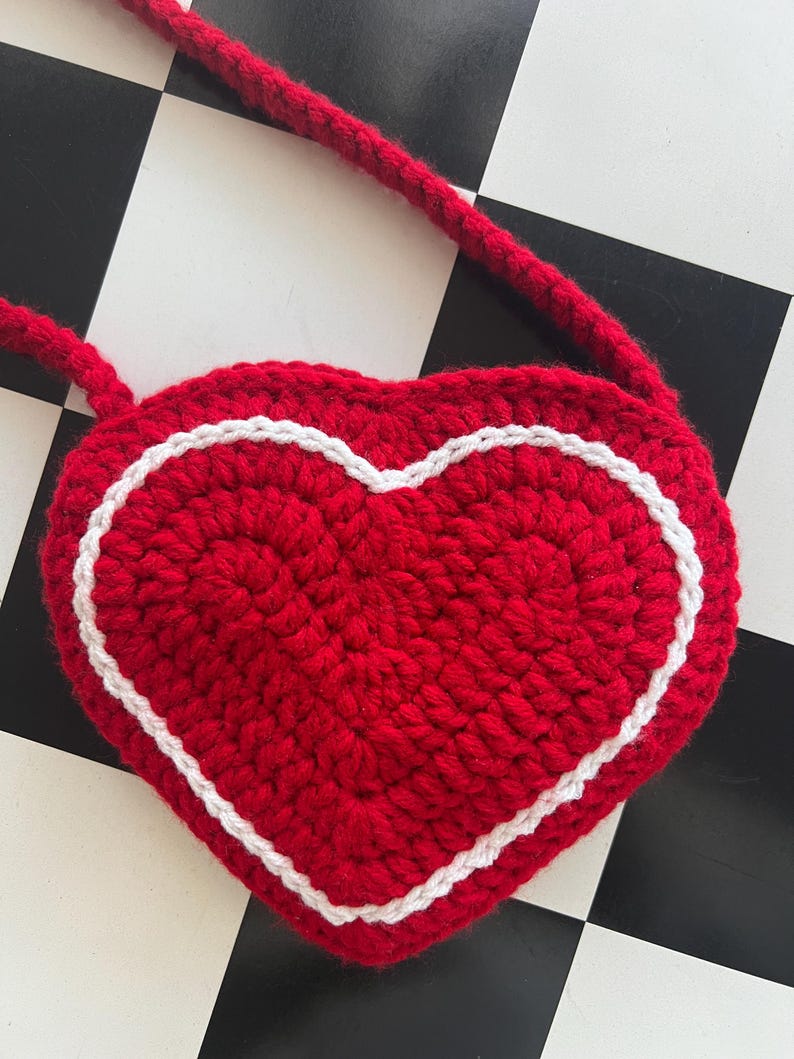 May include: A handmade, red crochet heart-shaped bag with a white border and a matching red strap. The bag is a vibrant red color, and the white border adds a contrasting detail. The bag is designed to be worn over the shoulder.