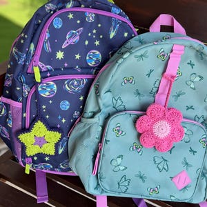 May include: Two backpacks, one dark blue with a space-themed print of planets and stars featuring a lime green crocheted star charm, and one light teal with a butterfly print featuring a pink crocheted flower charm, both with purple zippers.