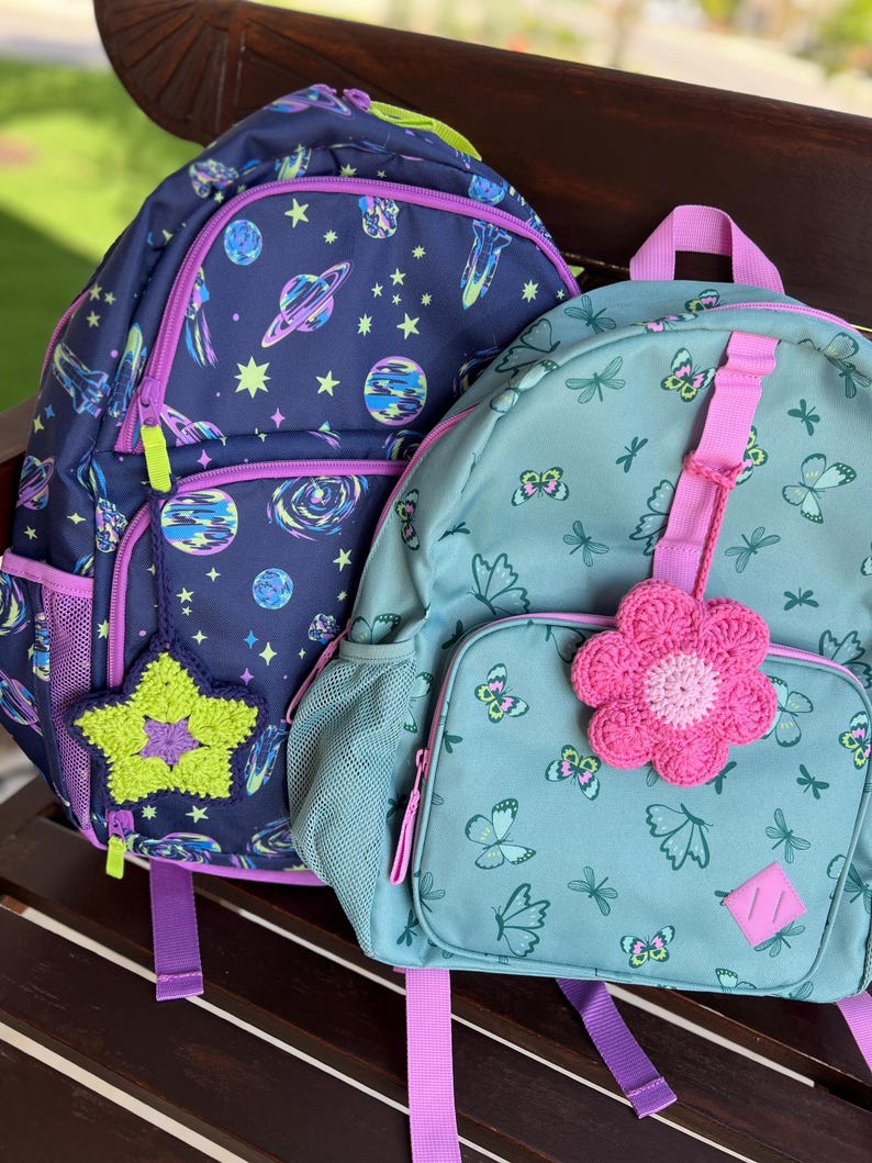 May include: Two backpacks, one dark blue with a space-themed print of planets and stars featuring a lime green crocheted star charm, and one light teal with a butterfly print featuring a pink crocheted flower charm, both with purple zippers.