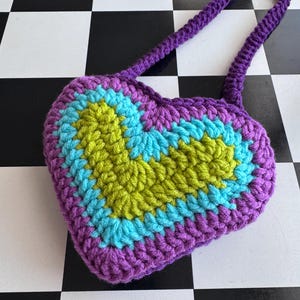 May include: A crocheted heart-shaped bag in shades of purple, blue, and green. The bag has a long, braided purple strap. The heart design features concentric stripes of color, with a green center and blue and purple borders. The bag is on a black and white checkered surface.