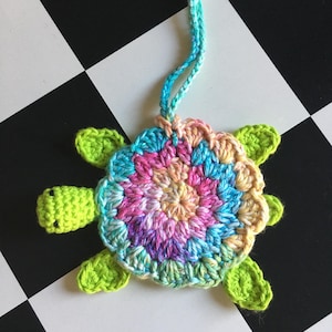 May include: A crocheted turtle ornament featuring a bright lime green head and four flippers. The shell is crafted from variegated yarn in shades of pink, blue, yellow, and purple, arranged in a circular pattern. A matching blue and green hanging loop is attached.