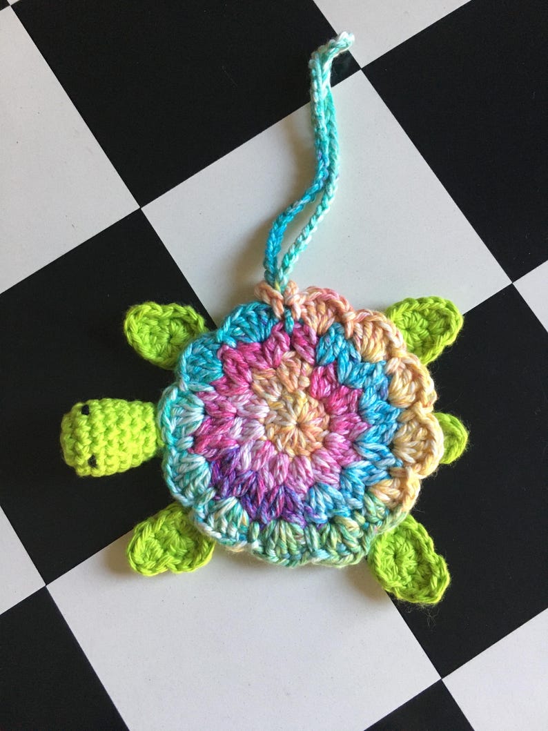 May include: A crocheted turtle ornament featuring a bright lime green head and four flippers. The shell is crafted from variegated yarn in shades of pink, blue, yellow, and purple, arranged in a circular pattern. A matching blue and green hanging loop is attached.