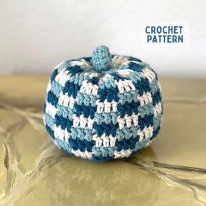 May include: A round, stuffed crochet pumpkin featuring a checkered pattern in shades of dark blue, light blue, and white. The pumpkin has a small, matching crocheted stem at the top center. A white rectangular label displays the text CROCHET PATTERN.