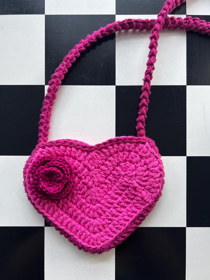 May include: A handmade, magenta-colored crochet heart-shaped purse with a matching strap. A crocheted rose is attached to the front of the purse. The purse is displayed on a black and white checkered surface.