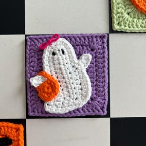 Op de afbeelding: Een vierkante gehaakte patch met een witte geest met twee zwarte ogen en een kleine roze strik op het hoofd. De geest houdt een feloranje pompoen vast. De hele figuur is gecentreerd op een effen paarse gehaakte vierkante achtergrond.