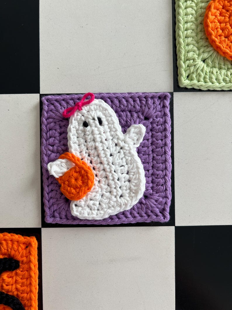 Op de afbeelding: Een vierkante gehaakte patch met een witte geest met twee zwarte ogen en een kleine roze strik op het hoofd. De geest houdt een feloranje pompoen vast. De hele figuur is gecentreerd op een effen paarse gehaakte vierkante achtergrond.