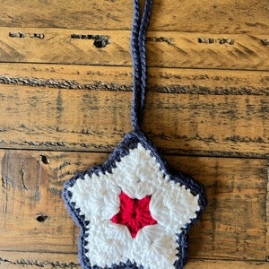 May include: A crocheted star ornament with a red center and white and blue accents. The star is hanging from a blue cord.