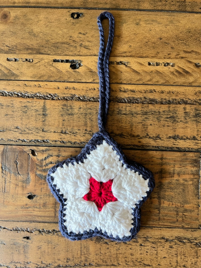 May include: A crocheted star ornament with a red center and white and blue accents. The star is hanging from a blue cord.