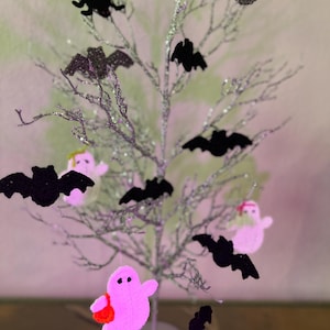 Op de afbeelding: Een zilveren glitterende decoratieve boom versierd met zwarte gehaakte vleermuis- en spinnenornamenten, naast roze gehaakte spookfiguren. De spoken hebben kleine zwarte ogen en dragen kleine oranje of groene tasjes aan de takken.