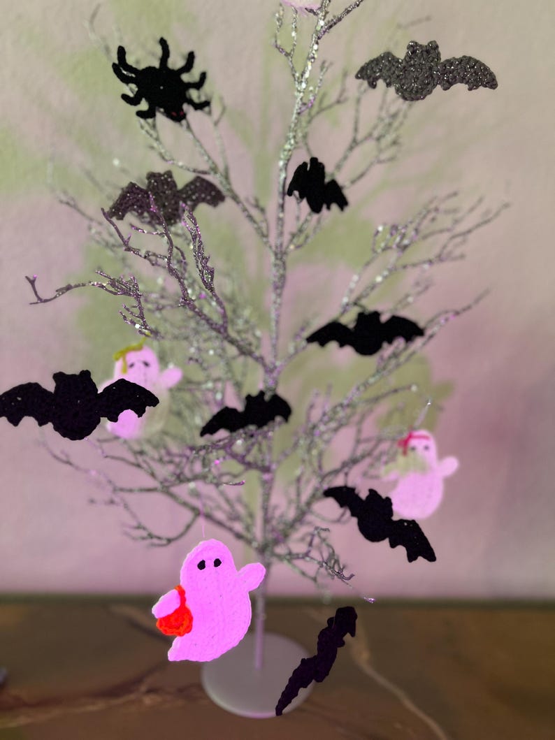 Op de afbeelding: Een zilveren glitterende decoratieve boom versierd met zwarte gehaakte vleermuis- en spinnenornamenten, naast roze gehaakte spookfiguren. De spoken hebben kleine zwarte ogen en dragen kleine oranje of groene tasjes aan de takken.