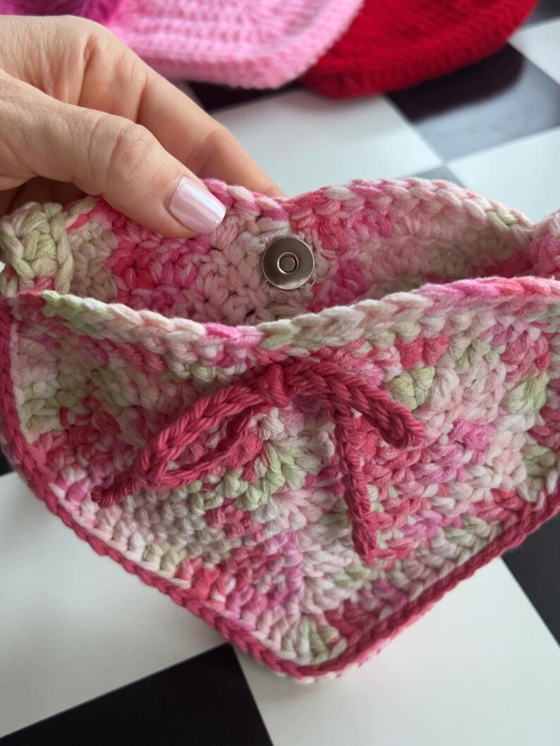 May include: A handmade crochet pouch in shades of pink and green, with a magnetic snap closure. The pouch features a drawstring closure and a heart-shaped design. The yarn has a variegated pattern, and the edges are finished with a darker pink yarn.