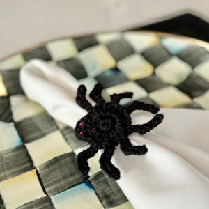 Op de afbeelding: Een zwarte gehaakte spinnen-servetring met acht dunne pootjes en twee kleine roze geborduurde oogjes. De spin is gemaakt van donker garen en is geplaatst rondom een gevouwen wit stoffen servet, wat zorgt voor een decoratief accent op tafel.