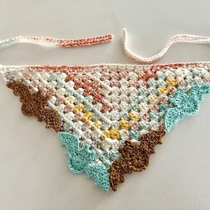 May include: A crocheted triangle scarf with a colorful geometric pattern. The scarf has a brown, pink, yellow, and blue color scheme. The edges of the scarf are scalloped and feature a bat design.