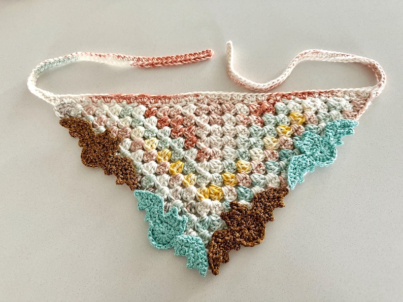 May include: A crocheted triangle scarf with a colorful geometric pattern. The scarf has a brown, pink, yellow, and blue color scheme. The edges of the scarf are scalloped and feature a bat design.
