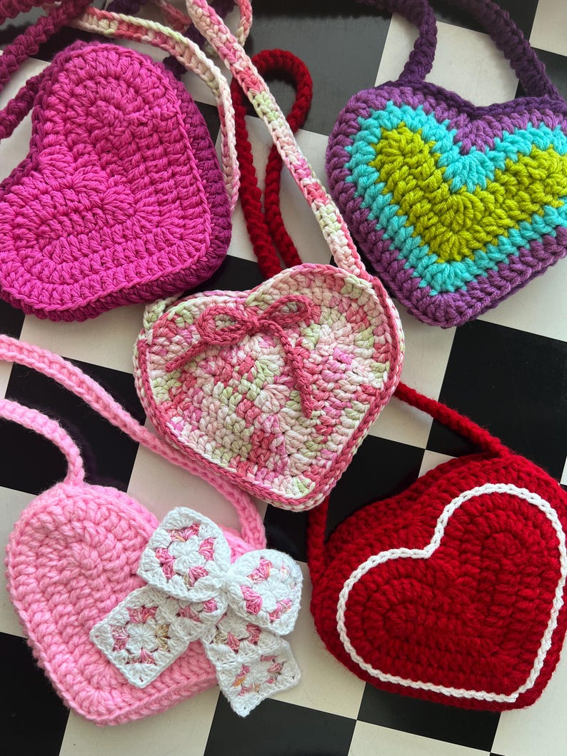 May include: A collection of handmade crochet heart-shaped bags in various colors, including pink, red, purple, and green. Some bags feature decorative bows and contrasting trim. The bags have straps for carrying.