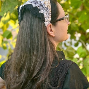 May include: A woman with long brown hair wearing a black crocheted headband with a white and gray pattern. The headband has a scalloped edge.