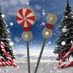 May include: Two decorative lollipop-shaped ornaments with silver stems. One has a red and white pinwheel design, the other has two white pearl-like spheres and a red disc. Candy canes are scattered in the snow.