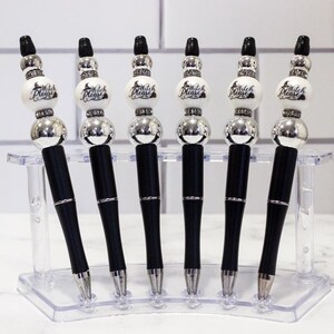 May include: Six black pens with silver accents and decorative tops. Each pen has a white bead with the words "Witch Please" printed on it. The pens are displayed in a clear plastic holder.