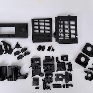 Op de afbeelding: Zwarte 3D-printer onderdelen, waaronder een rechthoekig onderdeel met de tekst "ORIGINAL PRUSA", diverse beugels en andere mechanische componenten. De onderdelen zijn gerangschikt op een witte ondergrond.