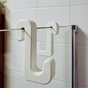 Op de afbeelding: Witte, U-vormige douchedeurhaken. De haken zijn gemaakt van een stevig, getextureerd materiaal en zijn ontworpen om over een glazen douchedeur te hangen. De haken worden getoond tegen een witte betegelde muur.